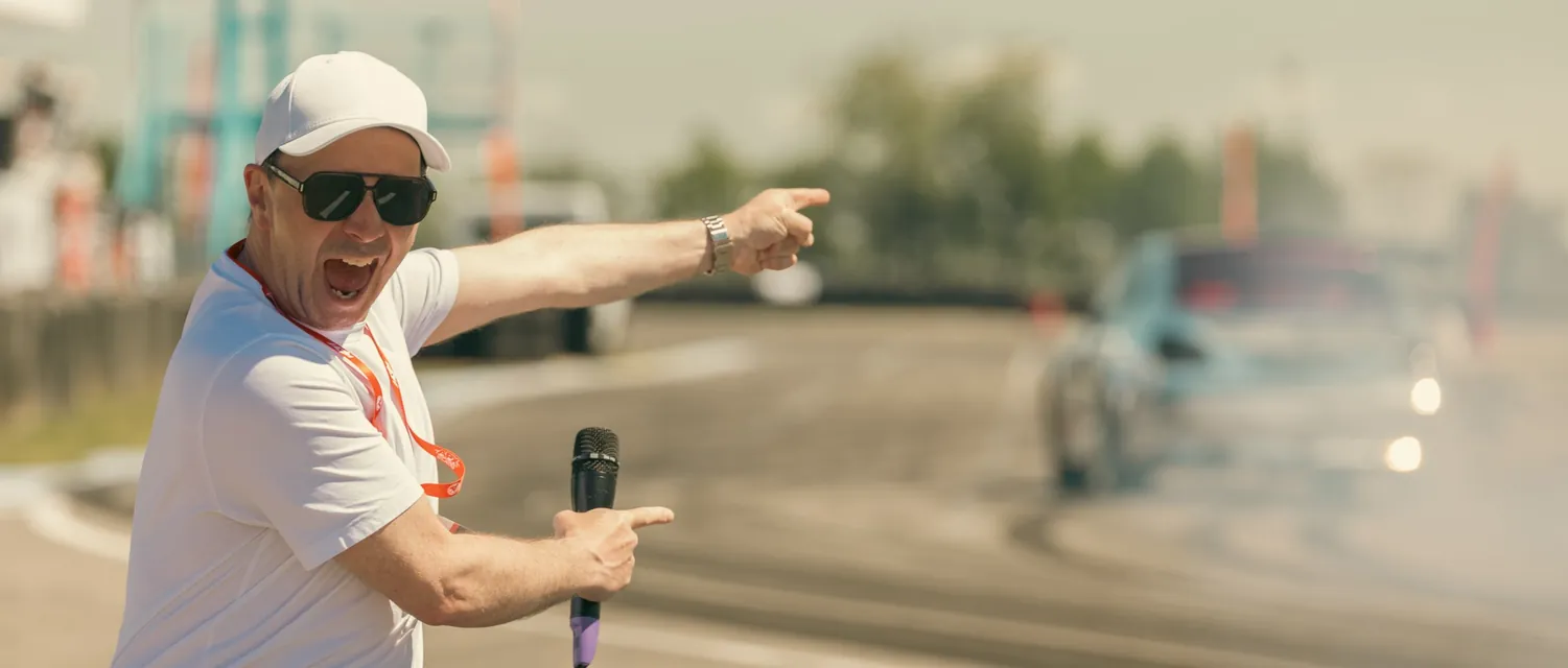 The announcer with a microphone points at a smoking car from the trackside