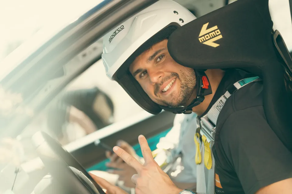 A smiling driver in a helmet in the race seat, making a victory sign