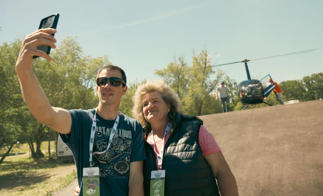 Two visitors take a selfie with a helicopter behind them by the track