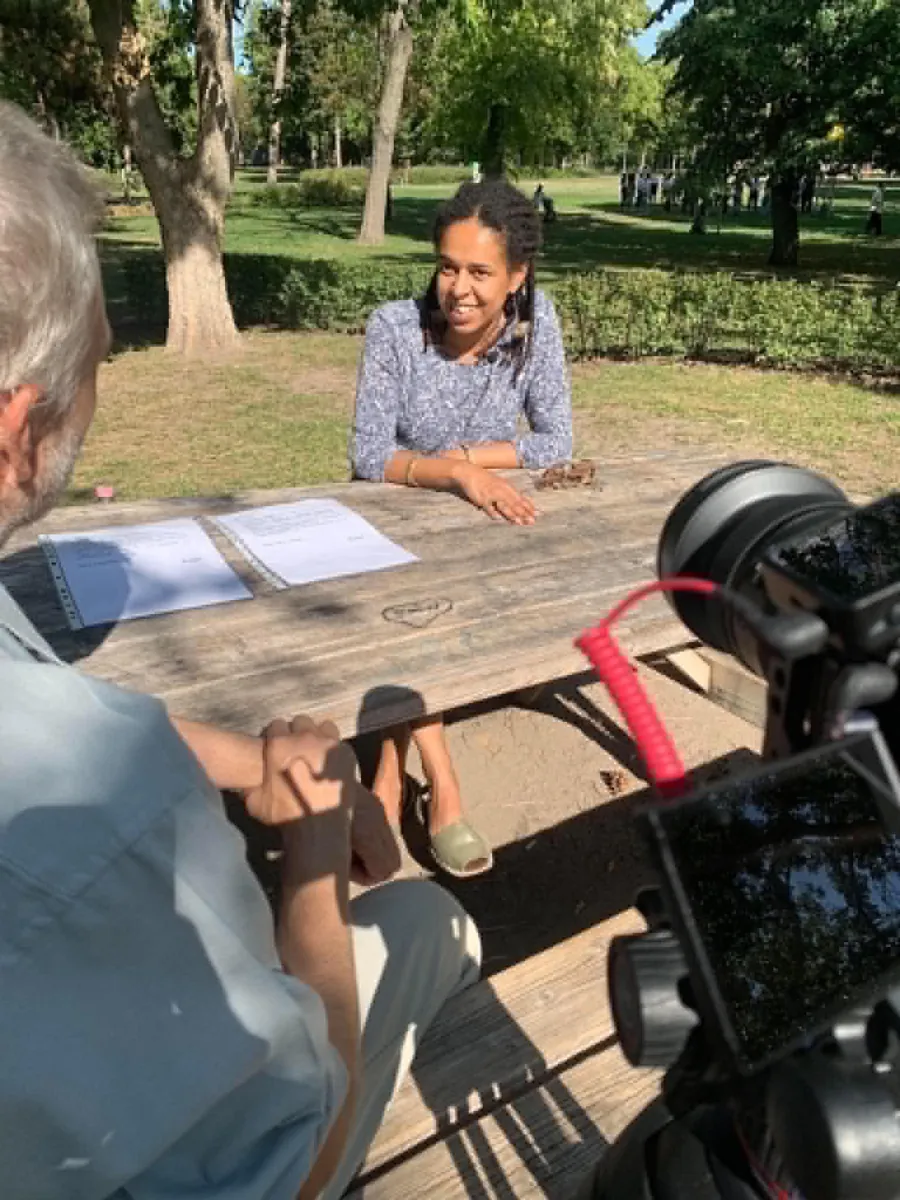 The first interview with Fatime Songoro in a park: Fatime smiles at a wooden table, with the camera and a crew member in front of her
