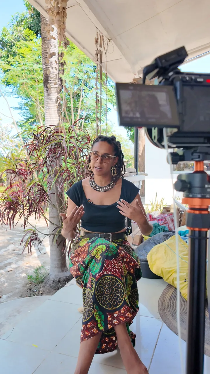 Fatime Songoro talking on a porch in a colourful African print skirt, with the filming camera and its flip-out monitor in the foreground