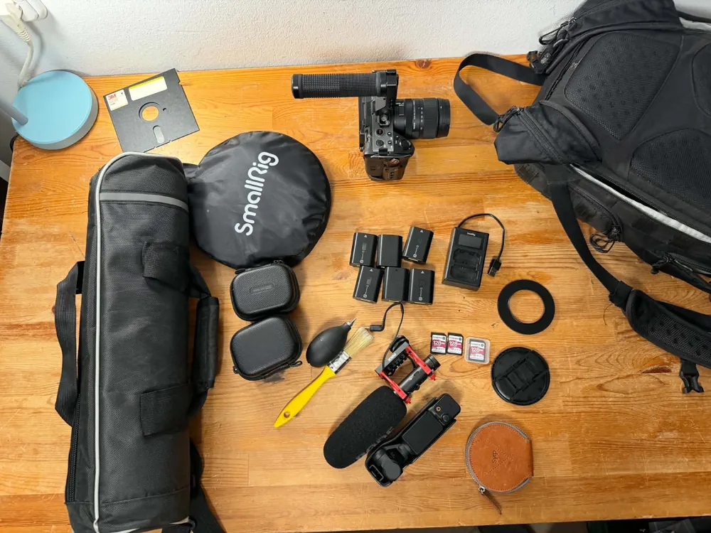 Filming kit laid out on a table: camera with a grip, microphone, six batteries, charger, memory cards, reflector and tripod bag