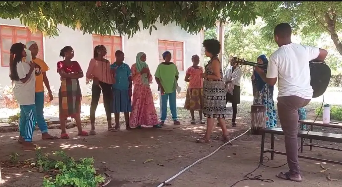 Outdoor music session in the shade of a tree: girls and women sing in a semicircle while a man plays the guitar