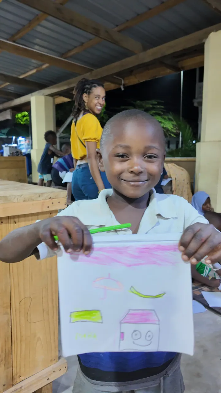 A smiling boy shows his drawing, with Fatime laughing behind him