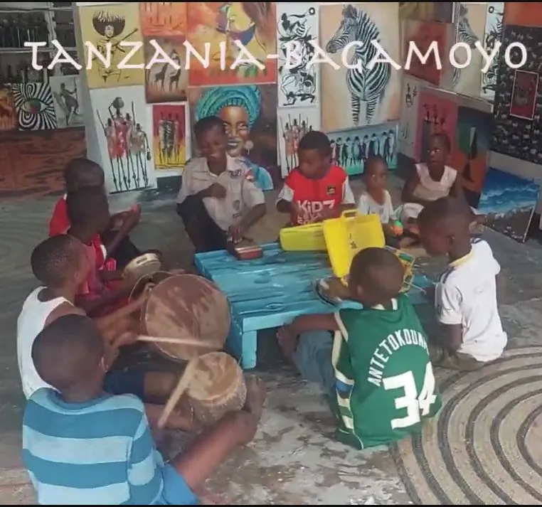 Children with drums around a blue table, in front of a wall of paintings with the caption Tanzania, Bagamoyo